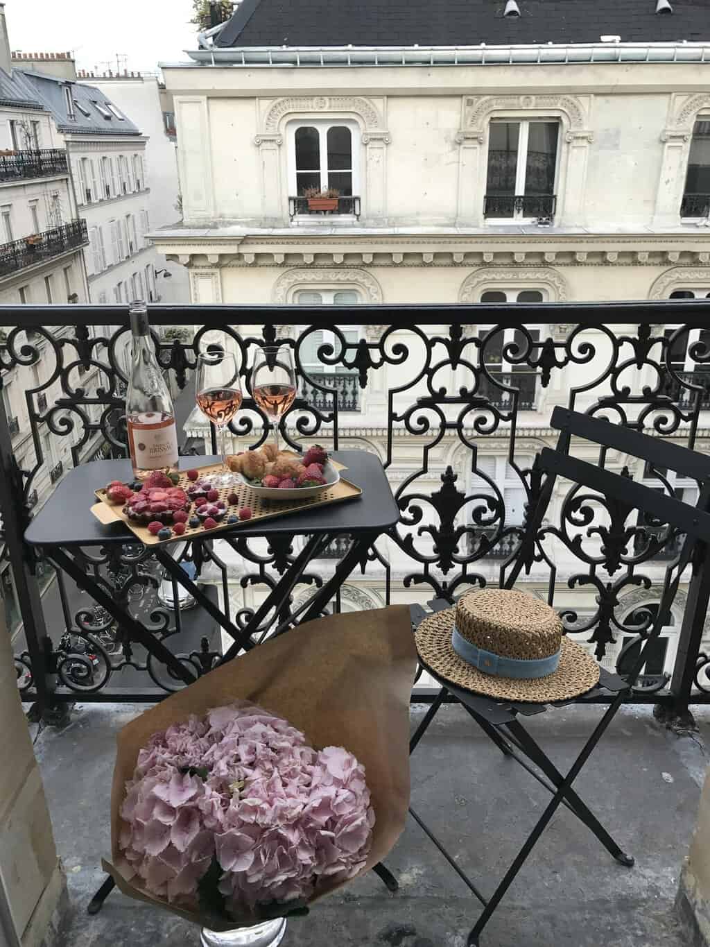 Cozy Parisian balcony setup with wine, snacks, and flowers on a sunny day.