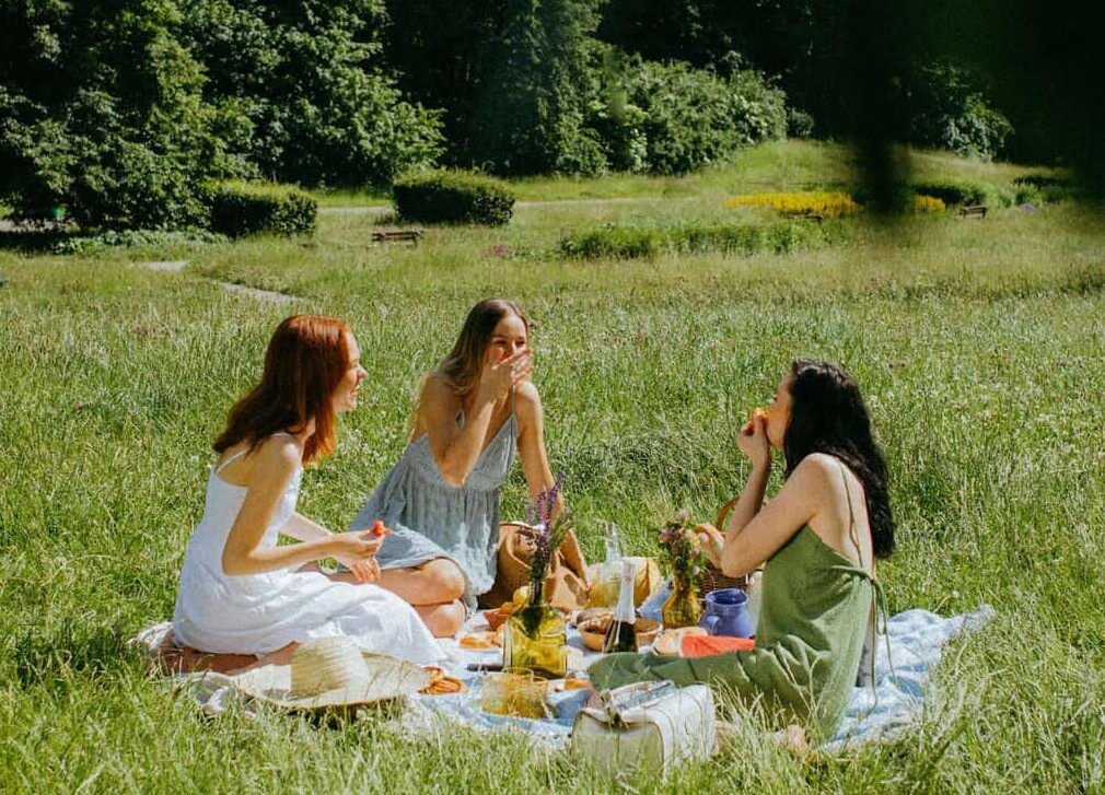 Three women enjoy a serene picnic in a sunny park with lush greenery.